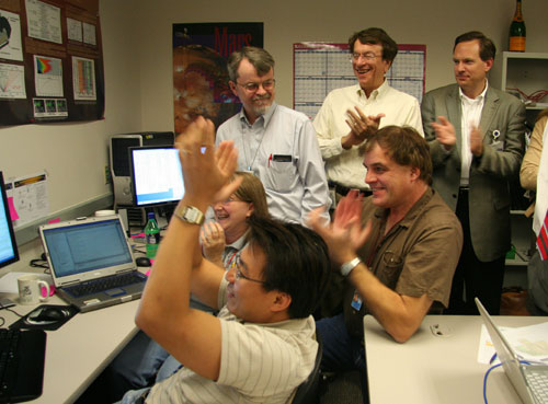 In this image, a small group of people clap while looking at computer screens that display data that indicates to them that their instrument is ready for operations.  Three of the people are seated and the rest of the people are gathered around them.