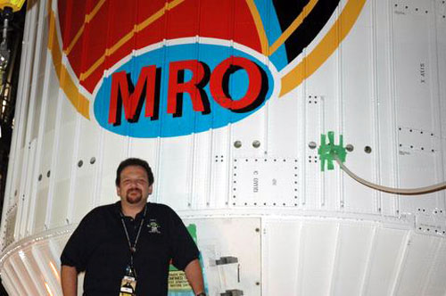 In this image, Howard Eisen, a Caucasian man in his thirties or forties with brown hair and a brown goatee stands in front of the large white protective fairing for the Mars Reconnaissance Orbiter.  The fairing is mostly white and its construction resembles siding for houses or corrugated tin.  The blue, yellow, red and black logo designed for the mission is partially visible.