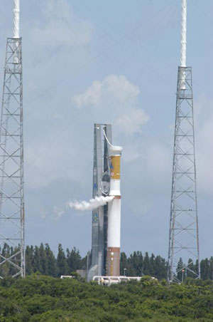 This image is a view of Launch Complex 41 at Cape Canaveral Air Force Station in Florida.  The large Atlas V rocket that will carry the Mars Reconnaissance Orbiter through Earth's atmosphere sits tall and majestic against a light blue sky blotted with very faint white clouds.  The lower stage of the rocket (closest to the ground) is half copper-colored and half white.  The upper stage, which is roughly half the size of the lower stage, is white and gold.  White smoke billows from the adaptor area where the stages meet as the 'wet dress rehearsal' is conducted.  To the left and right of the rocket sit two large towers that serve as lightning rods to protect the rocket and spacecraft from strikes.