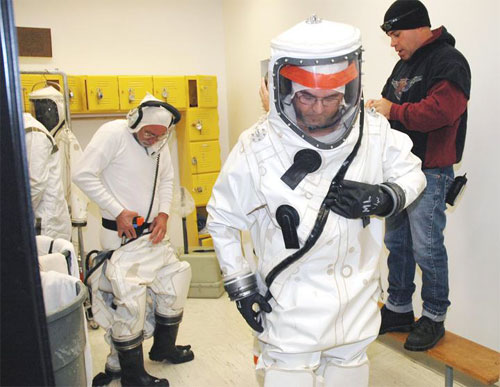 In this image, two men are in various stages of gearing up in suits that will protect them as they fuel the Deep Impact Spacecraft.  The suits are white with black accents.  These suits are distinctly different from regular cleanroom 'bunny suits.'  The gloves are large and black and cuffed by a silver protective band.  The boots, too, are large, black and secured to the leg by the same type of silver protective band.  The entire front of the large head gear is characterized by a clear, curved 'windshield' that, from the outside, reveals the wearer's entire head - not just his/her eyes as some cleanroom suits do.