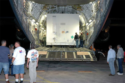 In this image, against the dark backdrop of the Florida night, the 'mouth'  of a large Air Force cargo plane is open, revealing one of two large white boxes containing the Mars Reconnaissance Orbiter.  The box is more than two times the height of the men standing in front of it, supervising its move off of the aircraft.