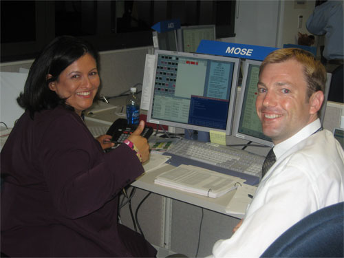 This photo shows Ruth Fragoso and Glen Havens turning from their shared computer screens that are spilling out lines and lines of spacecraft data to give a thumbs up and a smile to the camera.  Ruth is tan with shoulder-length, shiny black hair, and wears a dark plum-colored suit and a genuine smile with a cute dimple.  Glen has short blond hair, blue eyes, and a broad smile.  He wears a white dress shirt and a silver tie.  A blue sign with white letters, spelling 'MOSE,' sits on top of their computer screens.