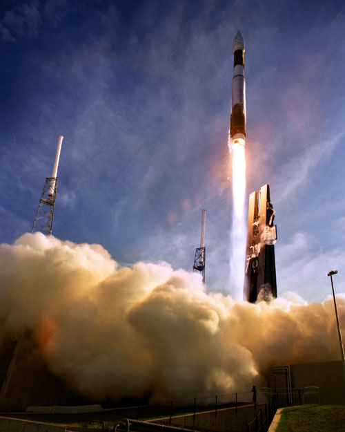 In this image, against a bright blue sky, an Atlas 5 rocket tears into the sky with a tail of fire leaving clouds of smoke in its wake.