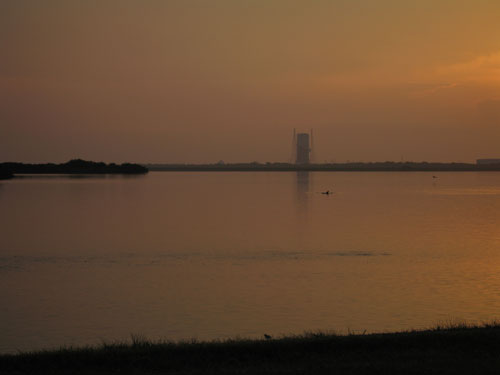 In this image, against a hazy but beautiful orange and pink sunrise, the dorsal fin of one of a pod of several dolphins is sticking up out of the calm water of the Banana River.  In the foreground of the image is the green, grassy area that surrounds the NASA Causeway on either side.  The Causeway is commonly used as the viewing site for launches from the Delta and Atlas launch pads.  In the center of the image, across the river, sits one of the Delta launch pads.