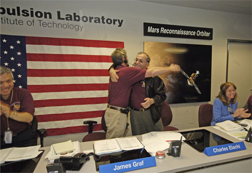 In this image, Graf, a middle-aged Caucasian man with brown hair and a goatee embraces Elachi, a middle-aged Lebanese-American man.  Both are wearing maroon-colored polo shirts with the mission logo on them.  Elachi also wears a black leather jacket and a wide smile.  They are surrounded by other NASA and JPL employees who are clapping.
