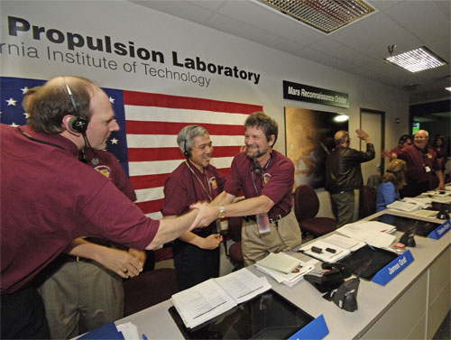 In this celebratory image, Graf, a middle-aged Caucasian man with brown hair and a goatee firmly shakes Dantzler's hand.  Dantzler is also a Caucasian man in his late thirties or early forties with thinning brown hair.  Li is a middle-aged Asian man.  They are in the large mission control room at JPL.  An American flag decorates the wall behind them.  Most team members are wearing maroon-colored polo shirts with the mission logo on them.