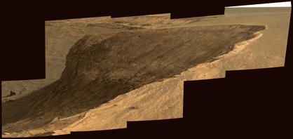This image shows a cliff of layered rocks beneath a sandy plateau. The landscape is brownish red.