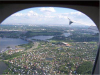 This picture was taken from inside an airplane as it was flying over Moscow at about 4,000 feet with a few clouds in the sky.  The left wing juts into view and the flaps are down.  The sun is shining on the community, which is speckled with hundreds of little farm-style houses, with lush green grass and trees surrounding all the buildings in the foreground.  In the distance, a wide, dark blue river snakes across the ground, with a large bridge crossing over the center of the river.  A few boats are docked near a road. In the far distance, thousands of buildings seemingly go on forever.