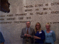 This picture is of Igor Mitrofanov, Nora Kelly and Cindy Schulz standing in front of a quote in Russian by Tsiolokovsky, who was a pioneer of rocketry in Russia.  Igor is a tall man with gray hair and glasses, wearing a grey blazer and red tie.  Nora is about 5'9, has long blond hair and is wearing glasses and a royal blue sweater.  Cindy is about 5'7 and wears a light blue sweater set.