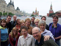 This picture is of fourteen adults smiling and huddling together in front of buildings in Red Square in Moscow.  The sky is grey and most people wear jackets.  In the background is a colorful church with eight onion-shaped domes called St. Basil's.  A large red brick building with a black and gold clock is to the right with a tall tower and a star on top.  To the left is an enormous white building that spans the width of the town square (about 200 feet) with scaffolding in front.