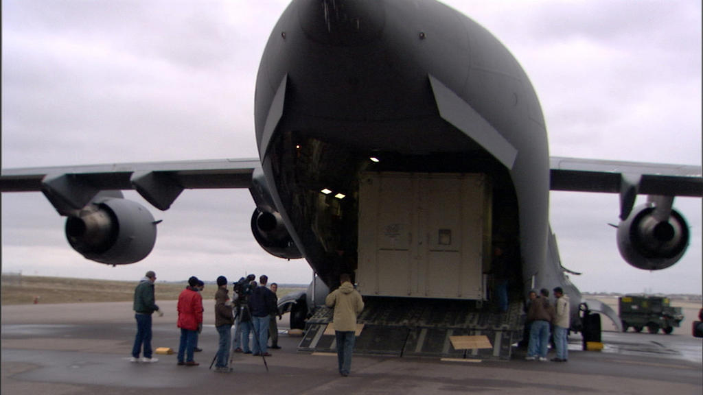 In this image, more than a dozen people stand, in winter coats, behind a huge, gray military C-17 cargo plane. From this vantage point, the plane resembles a huge shark with its mouth wide open. Inside the 'mouth' is one of the large boxes that contain the Mars Reconnaissance Orbiter. The wings of the plane jut out from both sides. The sky is cloud-covered.