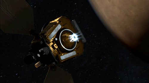 This image is a still animation of the boxy Mars Reconnaissance Orbiter spacecraft against the black background of space, dotted with stars.  Coming from the center of the boxy bus of the craft is white light that is meant to represent the firing of six main engines.  Extending from both sides of the spacecraft bus are the large solar panels that resemble two billboards.  The curve of Mars is on the right side of the image.  The planet is a muted rust color and its atmosphere is represented by a thin, ghostly blue.