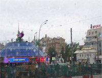 This image is a snapshot of Moscow from a bus on a rainy day.  A large, dome-like structure with blue, red, and pink neon signs houses a casino to the left in the picture; a fast-food restaurant sign blazes through the raindrops to the right; an old green and gold church peeks through the background; and a road construction zone complete with big, yellow Earth-mover trucks sits in the foreground.