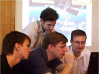 This image is of four young Russian engineers (each between 20 -25 years old) huddling together, intently looking at a laptop screen, discussing their next space mission.  Projected on a large screen behind the team is a picture of the space shuttle docked with the International Space Station orbiting the blue skies and oceans of Earth.