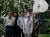 This picture is of six people smiling and standing in front of an old Venus spacecraft model that has been painted white and light blue.  They are in a green room full of large plants.  Vladislav wears a light grey oxford shirt with blue jeans and glasses, and has a crew-cut.  Elena has long brown hair and wears a large blue and black medallion necklace and a black suit.  Andrew O. wears glasses, a blue shirt and grey tie.  Serjey is about 5'5 and wears a light grey suit with a dark grey tie.  He has white hair and a white beard.  Andrew V. is about 6'1, has blond hair, wears glasses and a white shirt.  Sheri has short, light brown hair, and wears a dark silver, long jacket over black pants.