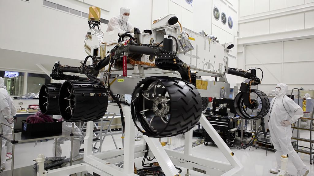 This image was taken in the clean room where the Curiosity rover is being assembled. It shows the rover, which is about the size of an SUV, hoisted on a white lift, with its black wheels suspended in the air. One engineer is on top of the hoist and is leaning over the rover body, while another is looking up on the ground floor to the right of the rover. Both engineers are wearing white "bunny suits" to keep them from contaminating any equipment.